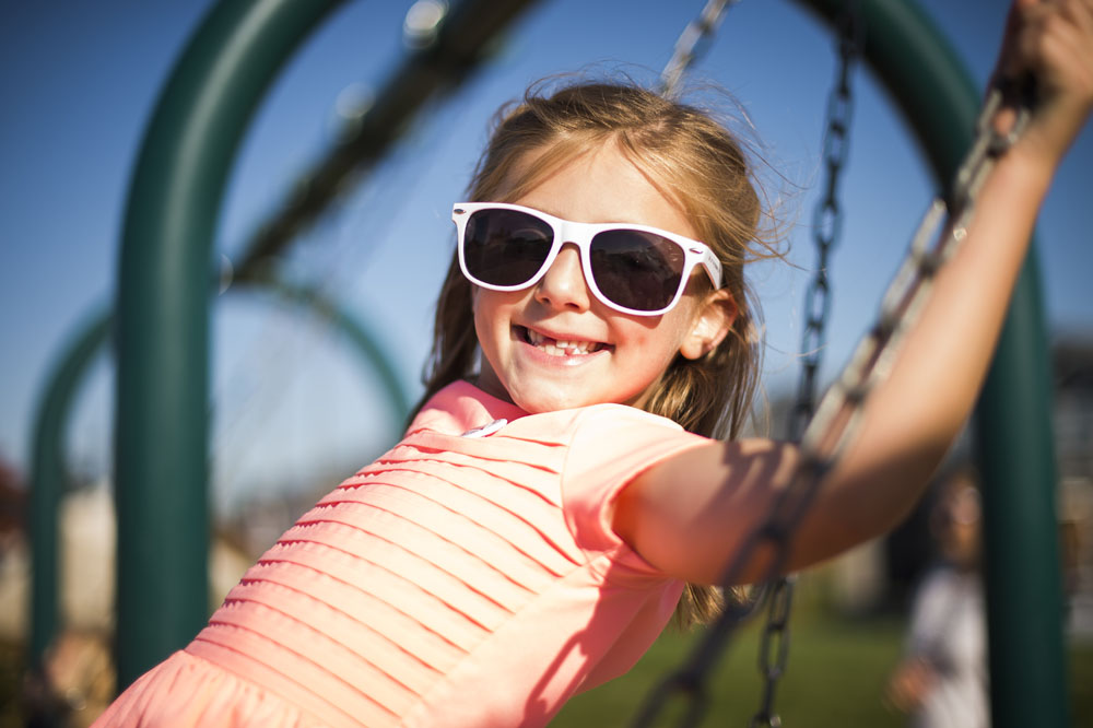 A closeup of a little girl with sunglasses smiling and holding on to a swing