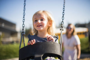 A closeup of a young girl smiling and sitting in the toddler-sized seat on a swingset