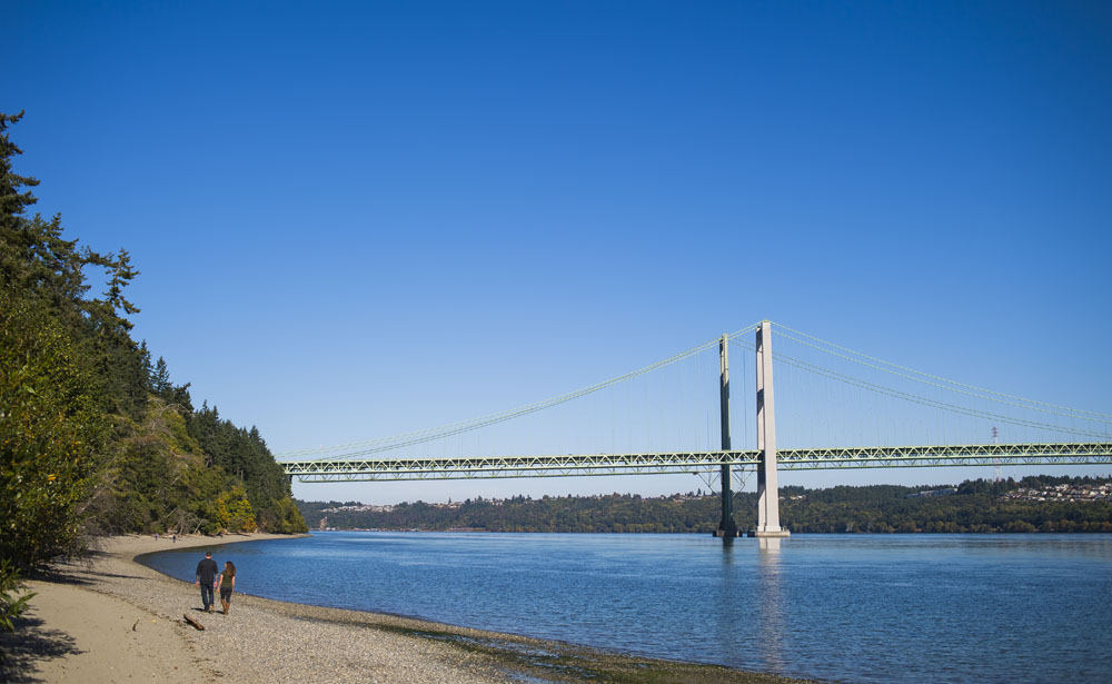 A couple walk along the beach of Narrows Park. The Tacoma Narrows Bridge is in the background.