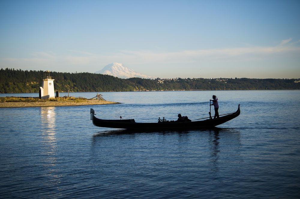 A side view of a man taking customers on a gondola ride in the harbor. Mount Rainier is in the background.