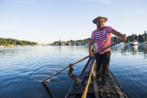 A closeup of a man smiling while holding a paddle to steer his gondola. An open sky is in the background.