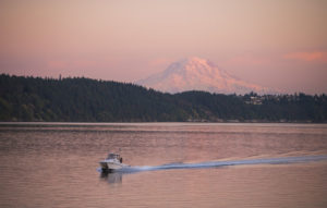 A motor boat goes through the harbor's empty water with Mount Rainier in the background. Both the water and the sky have turned pink from the sun setting.