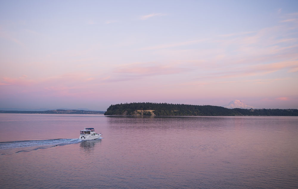 A motor boat goes through empty water. Some forested area is visible and Mount Rainier is in the background. The sky and water are pink from the sun setting.